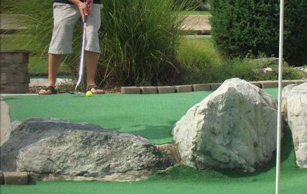 A person playing miniature golf. Picture shows their feet and them striking a golf ball with a putter
